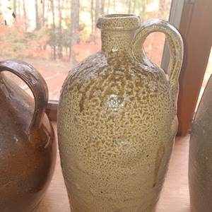 Puede incluir: Colección de jarras antiguas de gres con asas. Las jarras tienen una superficie moteada y texturizada en tonos beige y marrón. Las jarras se exhiben cerca de una ventana con vistas a los árboles.
