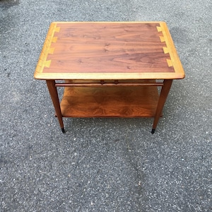 May include: A rectangular wooden side table featuring a dark brown walnut top with lighter wood dovetail joinery accents along the edges. The table includes a lower shelf and four tapered legs with black-tipped feet, constructed from wood.