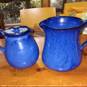 May include: Two blue ceramic pitchers with handles sit on a wooden surface. The pitcher on the left is smaller, with a lid. The pitcher on the right is larger, with a spout. Both have a glossy finish.