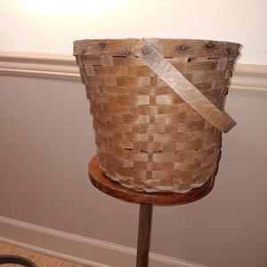 May include: A woven wooden basket with a wooden handle. The basket is light brown and sits on a small, round wooden stand. The basket has a cylindrical shape and is made of woven strips of wood.
