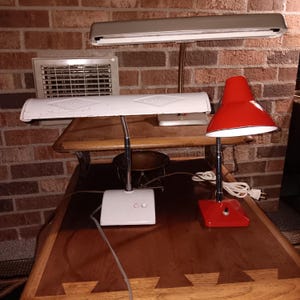 Puede incluir: Dos lámparas de escritorio vintage. Una es blanca con base rectangular y cuello flexible. La otra es roja con pantalla cónica y base cuadrada. Ambas lámparas están sobre un escritorio de madera.