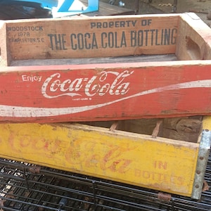 Puede incluir: Dos cajas de madera de Coca-Cola apiladas. La caja superior es roja con texto blanco que dice Enjoy Coca-Cola y Property of The Coca-Cola Bottling. La caja inferior es amarilla con texto descolorido Coca-Cola in Bottles. Tienen esquinas metálicas.