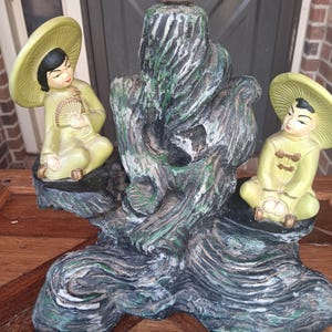 May include: A decorative figurine featuring two figures wearing light green conical hats and matching outfits, seated on a dark, textured, rock-like base. One figure holds a small fan, while the other sits with hands resting on knees.