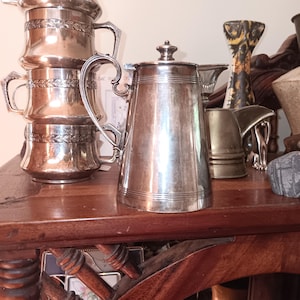 May include: A silver-toned metal coffee pot with a lid, a curved handle, and a spout, positioned next to a stack of three matching silver-toned metal cups featuring a decorative leaf-patterned band around the middle section of each cup.