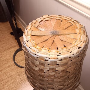 May include: A round, woven basket with a lid. The basket is made of light brown wood and has a cylindrical shape. The lid is made of the same material and has a cross-hatched design. The basket is likely used for storage.