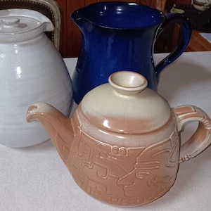 May include: Three ceramic kitchenware items: a white pitcher with a lid, a cobalt blue pitcher, and a brown teapot with a lid. The teapot has a handle and spout, and features a relief design.