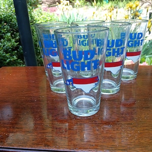Puede incluir: Un juego de seis vasos de pinta transparentes. Cada vaso presenta las palabras "Bud Light" en letras azules, con una franja roja y blanca y un contorno de Carolina del Norte. Los vasos están colocados sobre una superficie de madera.