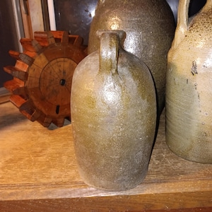 May include: A collection of antique stoneware jugs and a wooden gear. The jugs are various sizes and have a mottled, earthy color. One jug has a handle. The wooden gear is a warm brown color and has a rustic appearance.