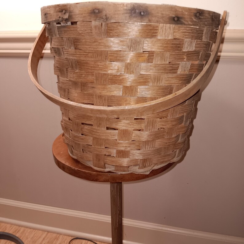 May include: A woven, light brown basket with a wooden handle. The basket is displayed on a small, round wooden stand with a tall, slender pole. The basket's weave is visible, and the handle arches over the top.