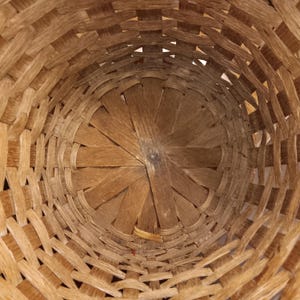 May include: A woven basket with a circular base and a cylindrical shape. The basket is made of light brown wooden strips, intricately woven to create a textured surface. The interior shows the woven pattern and the central base.