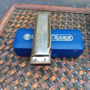 May include: A Hohner Blues Harp harmonica with a metallic gold and silver finish, featuring engraved text on the top plate. The instrument rests on a rectangular blue plastic storage case with white printed branding and the text Made in Germany.