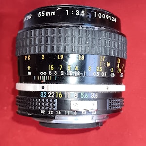 May include: A black Nikon 55mm camera lens with white and yellow markings. The lens has a ribbed focus ring and aperture settings from f/3.5 to f/32. The lens also has distance markings in feet and metres.