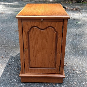 May include: A rectangular wooden end table with a medium brown oak finish. The piece features a single cabinet door with a decorative arched panel design, a flat top surface, and a small circular brass knob centered on the door frame.