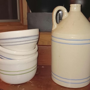 Op de afbeelding: Een verzameling vintage steengoed aardewerk. Een grote, gebroken witte kruik met een handvat en twee blauwe strepen. Gestapelde witte kommen met horizontale strepen in blauw, grijs en groen. Rustieke, boerderijstijl.