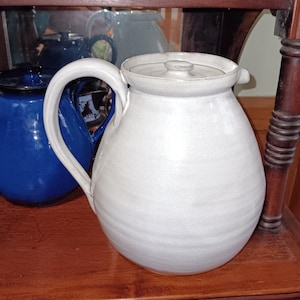 May include: A white ceramic pitcher with a curved handle and a lid. The pitcher has a rounded body and a spout. A blue ceramic pitcher is visible in the background. The pitchers are on a wooden shelf.