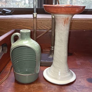 May include: Two ceramic pieces: a light green jug with a handle and a large opening, and a tall, slender vase with a wide, reddish-brown top. The vase has a light gray and white body. Both are on a wooden surface.
