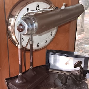 Może przedstawiać: Antyczna brązowa lampa biurkowa z cylindrycznym kloszem stoi przed dużym zegarem. Zegar ma białą tarczę z czarnymi cyframi. Na biurku znajduje się również mały czarny zegar i metalowy dziurkacz.