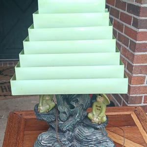 May include: A vintage table lamp with a tiered, light green shade. The base features a sculpted, dark green tree trunk design with two seated figures in yellow-green robes. The lamp sits on a wooden table.