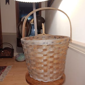 May include: A large, woven basket with a wooden handle. The basket is made of light brown wood and has a rounded shape. It is sitting on a small, round wooden table. The basket is likely used for storage or decoration.