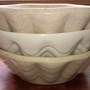 Puede incluir: Tres cuencos de cerámica apilados. El cuenco superior es de color beige moteado, el cuenco del medio es blanco y el cuenco inferior es de color beige claro. Todos los cuencos tienen un borde ondulado y festoneado.