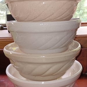 May include: Four stacked ceramic mixing bowls in varying shades of cream and beige. Each bowl has a textured, braided design around the exterior. The bowls are nested, with the largest at the bottom and the smallest at the top.
