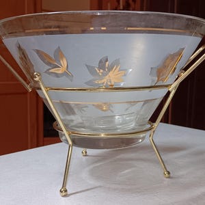 以下が含まれることがあります： 金色の葉のデザインが施された、つや消しブルーのバンドが付いた透明なガラス製のサービングボウル。金色の金属製のスタンドにセットされています。ボウルは幅広で開いた上部と、3本足のスタンドで支えられた小さなベースを備えています。