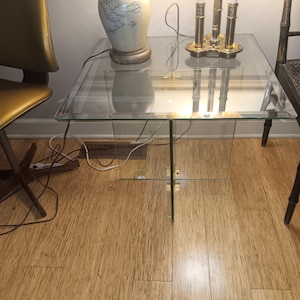 May include: A square glass side table with a clear top and legs. The table is supported by four glass panels that intersect at the centre. A gold-coloured metal accent is at the intersection of the legs. The table is on a wooden floor.
