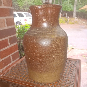 May include: A tall, cylindrical stoneware jug with a two-tone brown glaze. The upper portion features a smooth, dark brown finish, while the lower section transitions into a lighter, speckled tan hue. It has a wide mouth and horizontal ridges.