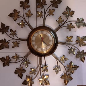 Pode incluir: Um relógio de parede decorativo com mostrador e ponteiros dourados, emoldurado em um círculo marrom escuro. O relógio é rodeado por um design metálico de folhas e galhos, criando uma estética inspirada na natureza.