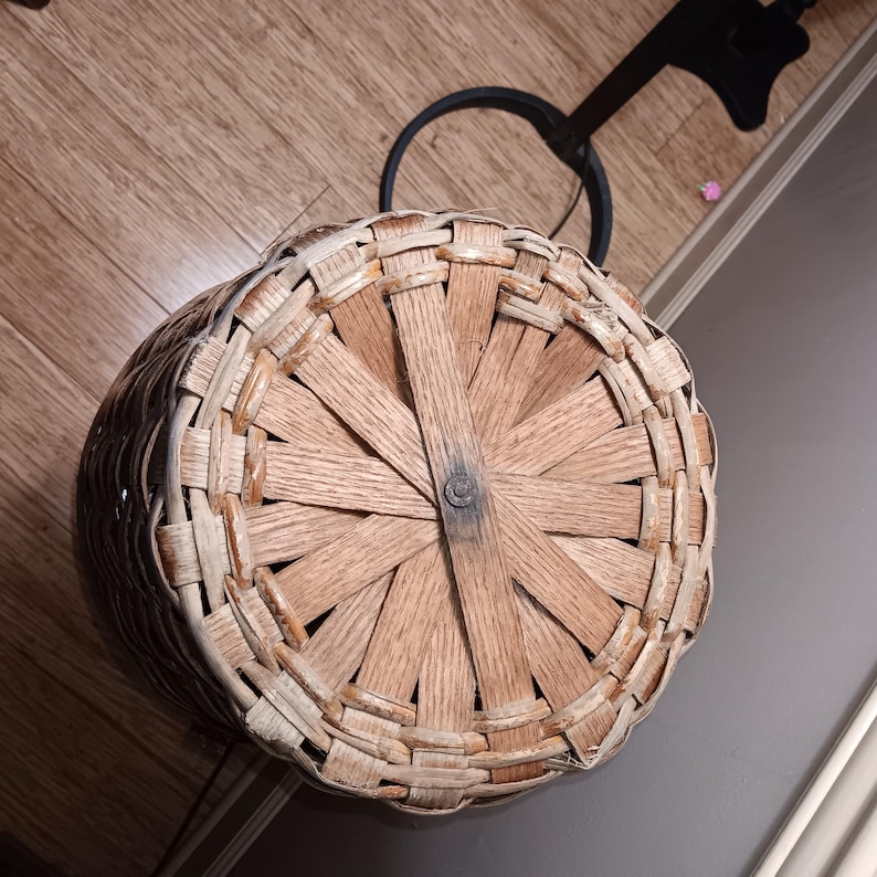 May include: A woven basket with a round base and a handle. The basket is made of light brown and beige woven material. The base is constructed with wooden slats radiating from a central point.