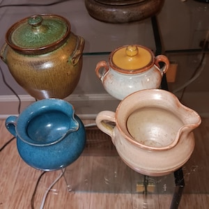 May include: A collection of handmade ceramic pottery pieces in various shapes and colors. Includes a lidded jar with a green lid, a small sugar bowl with an orange lid, a blue pitcher, and a beige pitcher. The pottery has a rustic, earthy aesthetic.