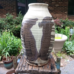 May include: A tall, cylindrical ceramic vase with a cream-colored base and horizontal ribbed texture. The exterior features two large, dark brown, abstract brushstroke patterns that mirror each other, creating a central negative space.