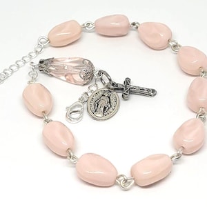 May include: A light pink beaded bracelet with silver-tone metal accents. The bracelet features oval-shaped beads, a cross charm, a religious medal, and a decorative glass bead. The bracelet has an adjustable chain.