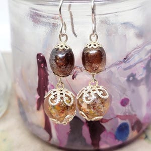 May include: A pair of dangle earrings with silver-colored hooks. Each earring features a dark brown, oval-shaped bead above a clear, round bead filled with colorful flecks. The beads are connected by silver-colored filigree caps.
