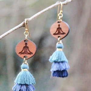 Puede incluir: Un par de pendientes de madera con la silueta de una persona en una postura de yoga. Los pendientes tienen un borla azul y turquesa.