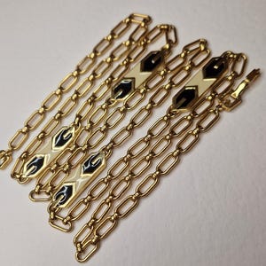 Puede incluir: Un collar de cadena de oro con detalles en esmalte negro y blanco. La cadena está hecha de eslabones ovalados y los detalles son formas geométricas.