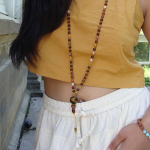 Puede incluir: Un collar largo de cuentas con un colgante. El collar presenta una variedad de cuentas de tonos tierra, incluyendo marrón, rojo y amarillo. El colgante es una cuenta más grande y redonda con un patrón de ojo de tigre. El collar se lleva sobre un top color mostaza.