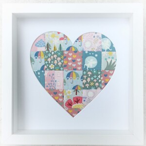 May include: A framed artwork featuring a heart shape. The heart is made from small fabric squares, each with a different pattern including umbrellas, houses, and flowers. The frame is white, offering a clean, contemporary aesthetic.