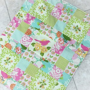 May include: A patchwork fabric with a green, pink and blue floral pattern. The fabric features a repeating pattern of squares with different floral designs.