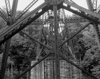 May include: Black and white photo of a metal bridge structure. The bridge is made of steel beams and supports. The bridge is over a body of water. The photo is taken from underneath the bridge.