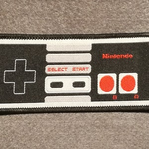 Peut inclure: Un patch brodé noir et blanc représentant une manette de jeu Nintendo NES avec le mot "Nintendo" en texte blanc. Le patch a une bordure blanche et des coutures noires.