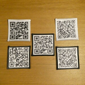 May include: Five black and white square patches with a QR code design. The patches are sewn onto a light brown wooden surface.