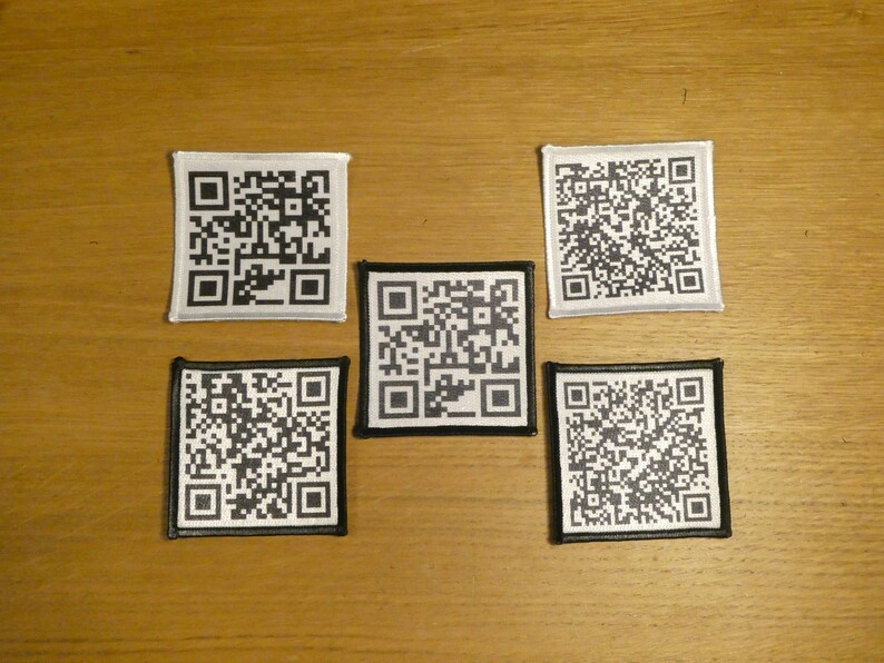 May include: Five black and white square patches with a QR code design. The patches are sewn onto a light brown wooden surface.