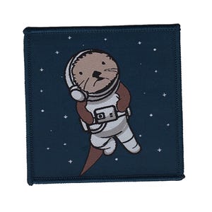 Może przedstawiać: Niebieska łatka z białą obwódką przedstawia kreskówkową wydrę w białym kostiumie astronauty z brązowym hełmem i słuchawkami. Wydra trzyma aparat i unosi się w kosmosie z gwiazdami w tle.