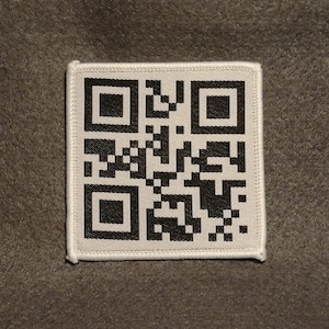 Op de afbeelding: Een witte stoffen patch met een zwart-witte QR-code erop gedrukt. De patch heeft een witte rand met een gestikte rand.