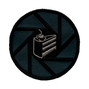 May include: A black and grey embroidered patch featuring a camera lens aperture design and a grey and black cake with a cherry on top.