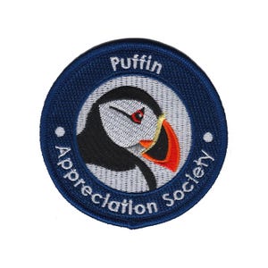 Può includere: Toppa rotonda ricamata con bordo blu navy e la scritta "Puffin Appreciation Society". La toppa presenta un'illustrazione di un pulcinella di mare con testa nera, faccia bianca e becco rosso, arancione e giallo. Sfondo bianco.