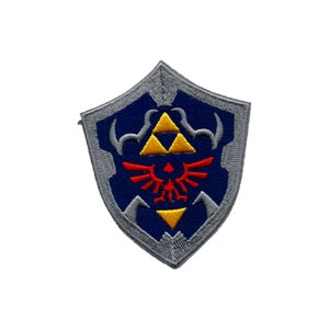 Op de afbeelding: Een blauwe en grijze geborduurde patch met het iconische Triforce-symbool uit de videogame-serie The Legend of Zelda. De Triforce wordt omringd door een gestileerd gevleugeld ontwerp en een grijze schildvorm.