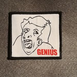 Op de afbeelding: Witte geborduurde patch met een zwarte lijntekening van een persoon met zijn mond open en het woord "GENIUS" in rode letters.