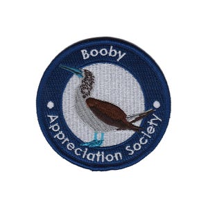 May include: Circular embroidered patch with a navy blue border. The patch features a blue-footed booby bird illustration within a white circle. Text reads "Booby Appreciation Society". The bird is depicted in brown, white, and blue.